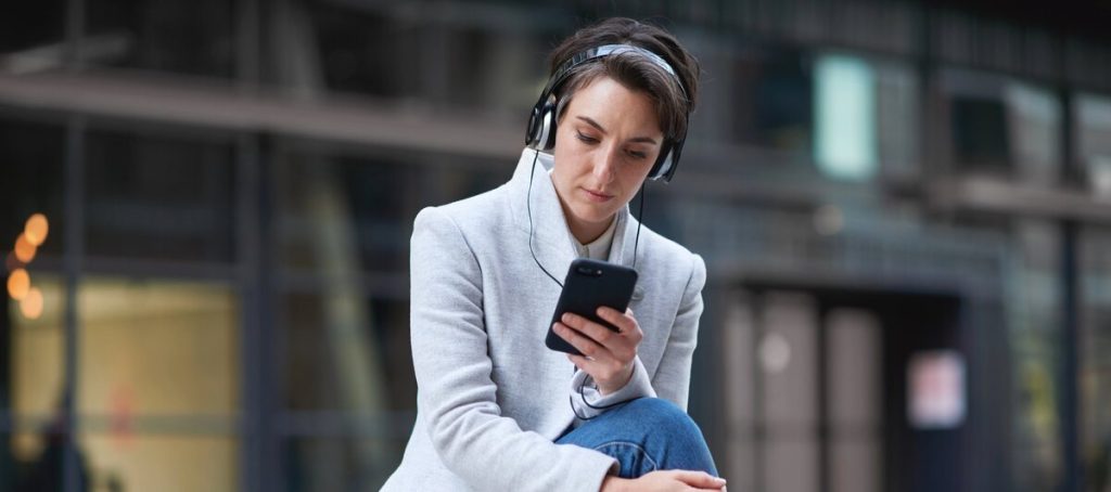 Mujer con auriculares sentada en la calle consulta un podcast en su móvil
