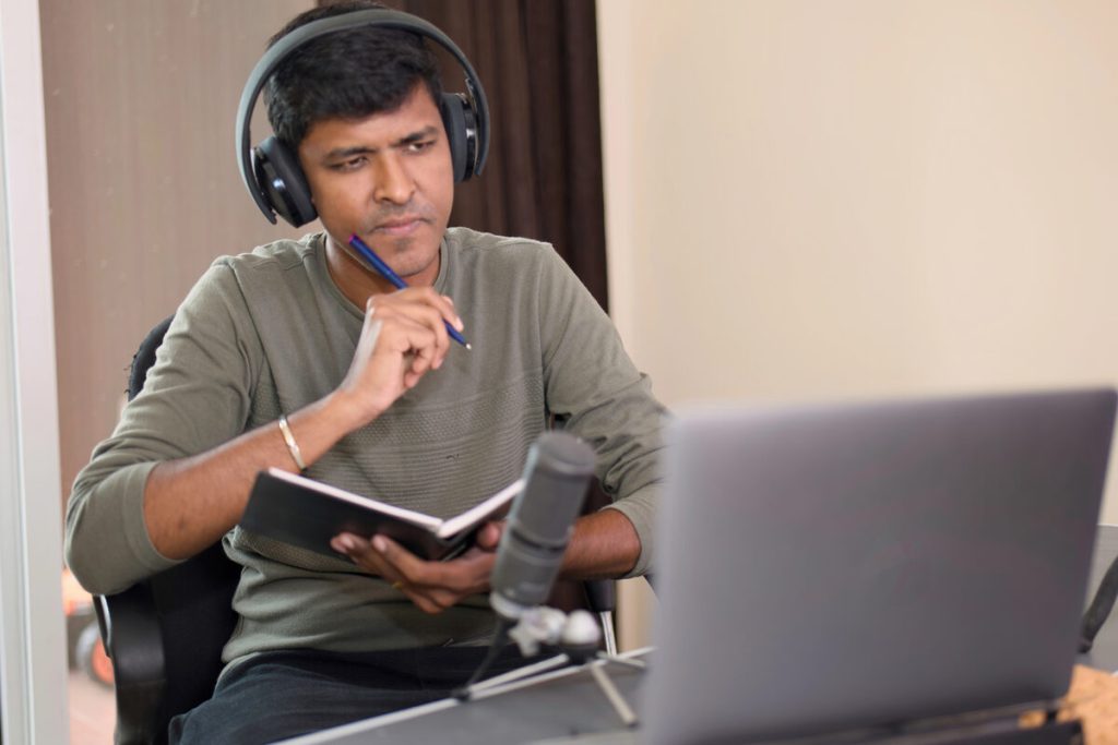 Podcaster con auriculares toma notas mientras revisa la propuesta de un invitado en el portátil